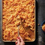 A hand scooping a spoonful of baked macaroni and cheese out of a sheet pan, seen from above.